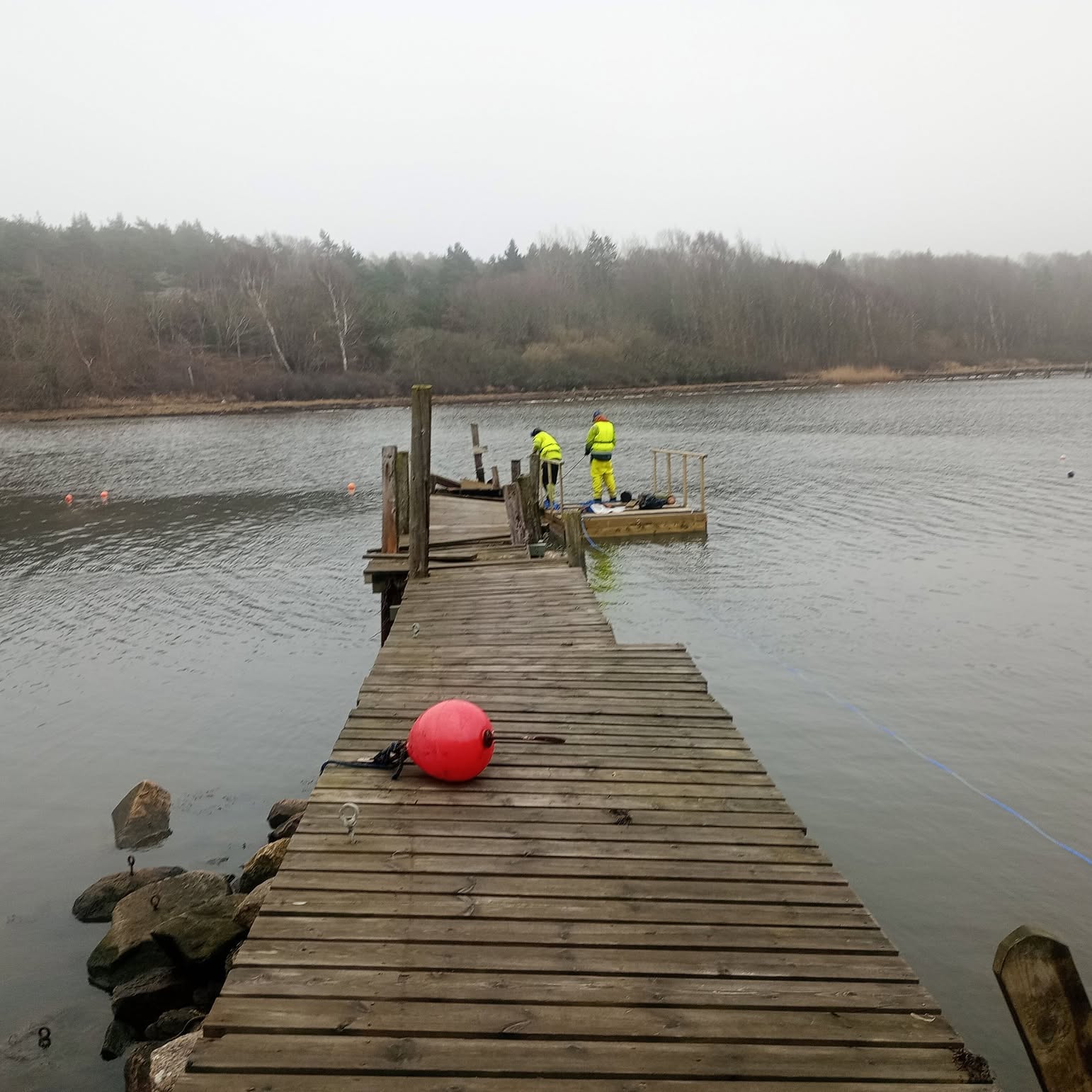 Två medarbetare i varselkläder renoverar en brygga vid havet en dimmig dag