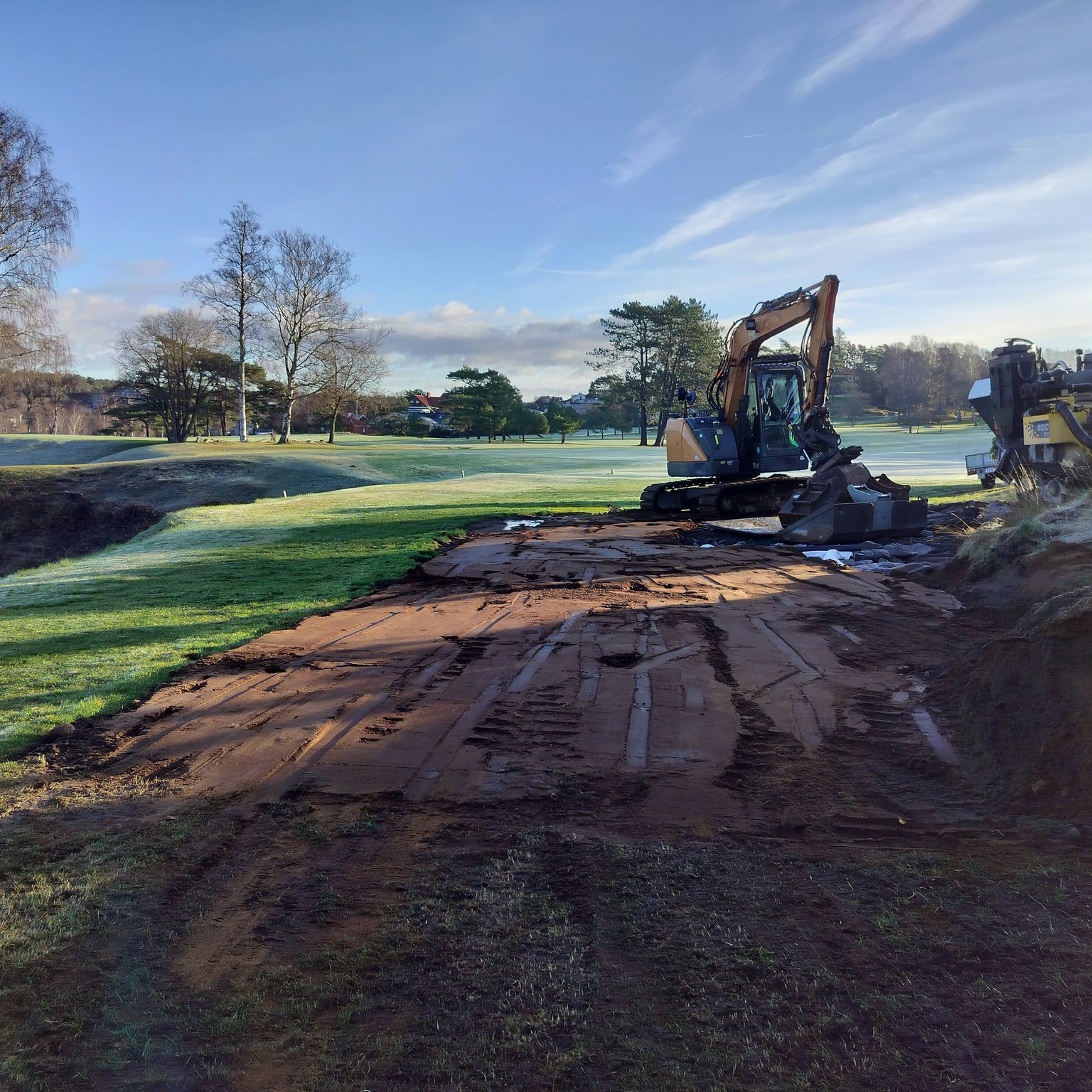 Grävmaskin och borrigg vid schakt- och dräneringsarbete på golfbana i morgonljus